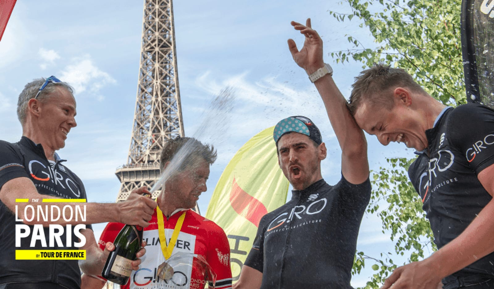 Four cyclists celebrate by spraying champagne near the Eiffel Tower. One cyclist in a red and white jersey holds a bottle while three others in black jerseys laugh and cheer. A "London to Paris" sign is visible in the foreground. The sky is clear.