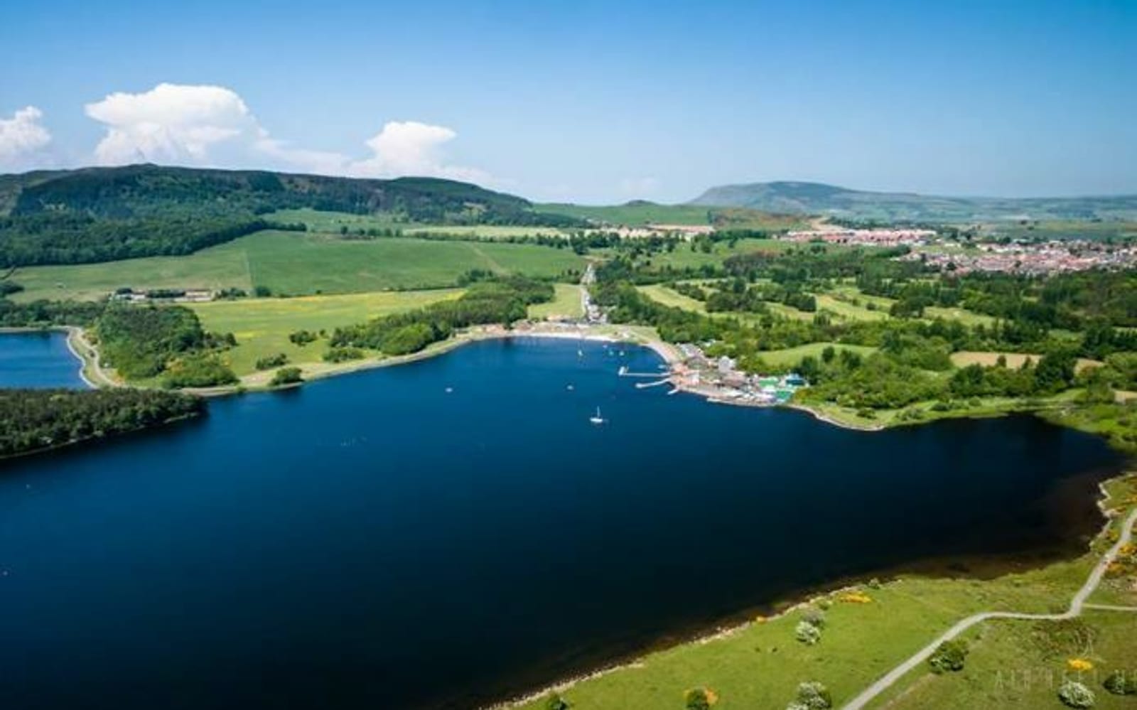 Aerial view of a tranquil lake surrounded by lush green fields, trees, and small hills. On the lake's shore, there are several docks and boats, with a cluster of buildings nearby. A small town is visible in the distance, set against a backdrop of rolling hills and blue sky.