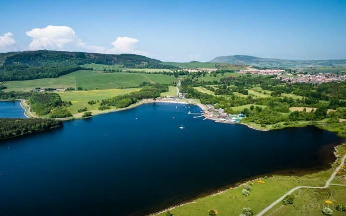 Aerial view of a tranquil lake surrounded by lush green fields, trees, and small hills. On the lake's shore, there are several docks and boats, with a cluster of buildings nearby. A small town is visible in the distance, set against a backdrop of rolling hills and blue sky.