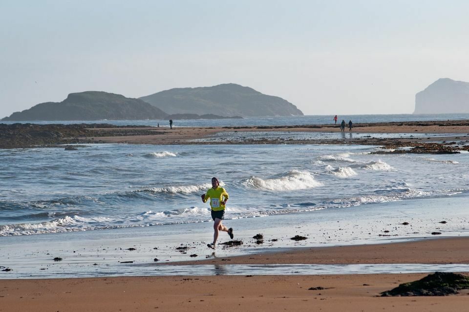 A person in a yellow shirt runs along a sandy beach with gentle waves in the background. Rocky outcrops and distant islands are visible in the sea. A few other people are walking along the shoreline. The sky is clear, indicating a bright, sunny day.