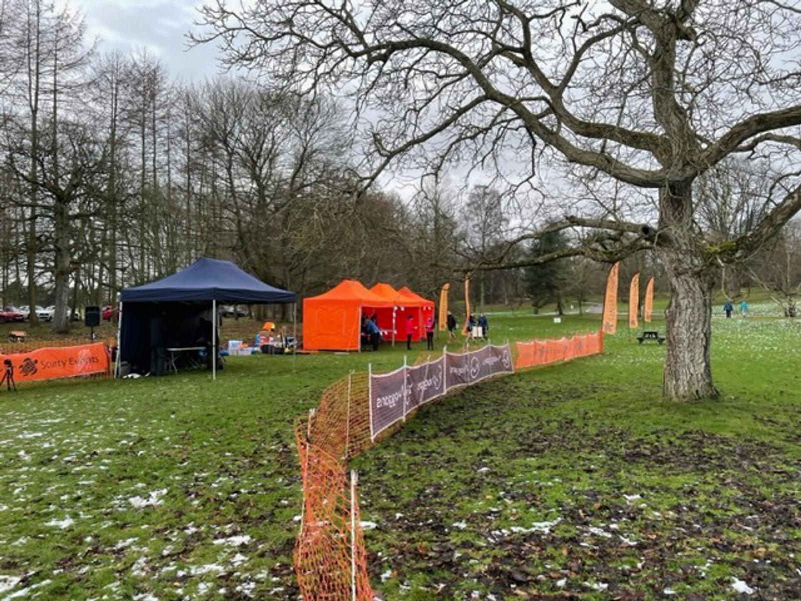 Outdoor event setup in a park with bare trees. The scene includes blue and orange tents, orange fencing, and promotional banners. Some snow patches are visible on the grass. People are gathered near the tents, engaging in event activities.