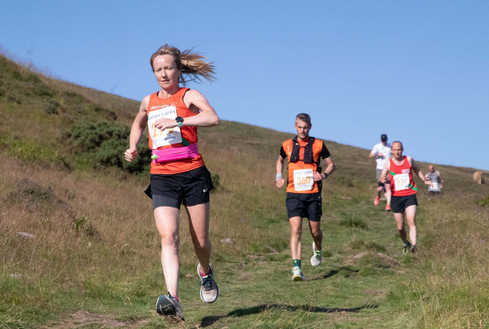 Three runners are participating in a trail race on a grassy hillside under a clear blue sky. The woman in front is wearing a red tank top with a pink band and black shorts, followed by two men, one in a black and orange running vest, and another in white.