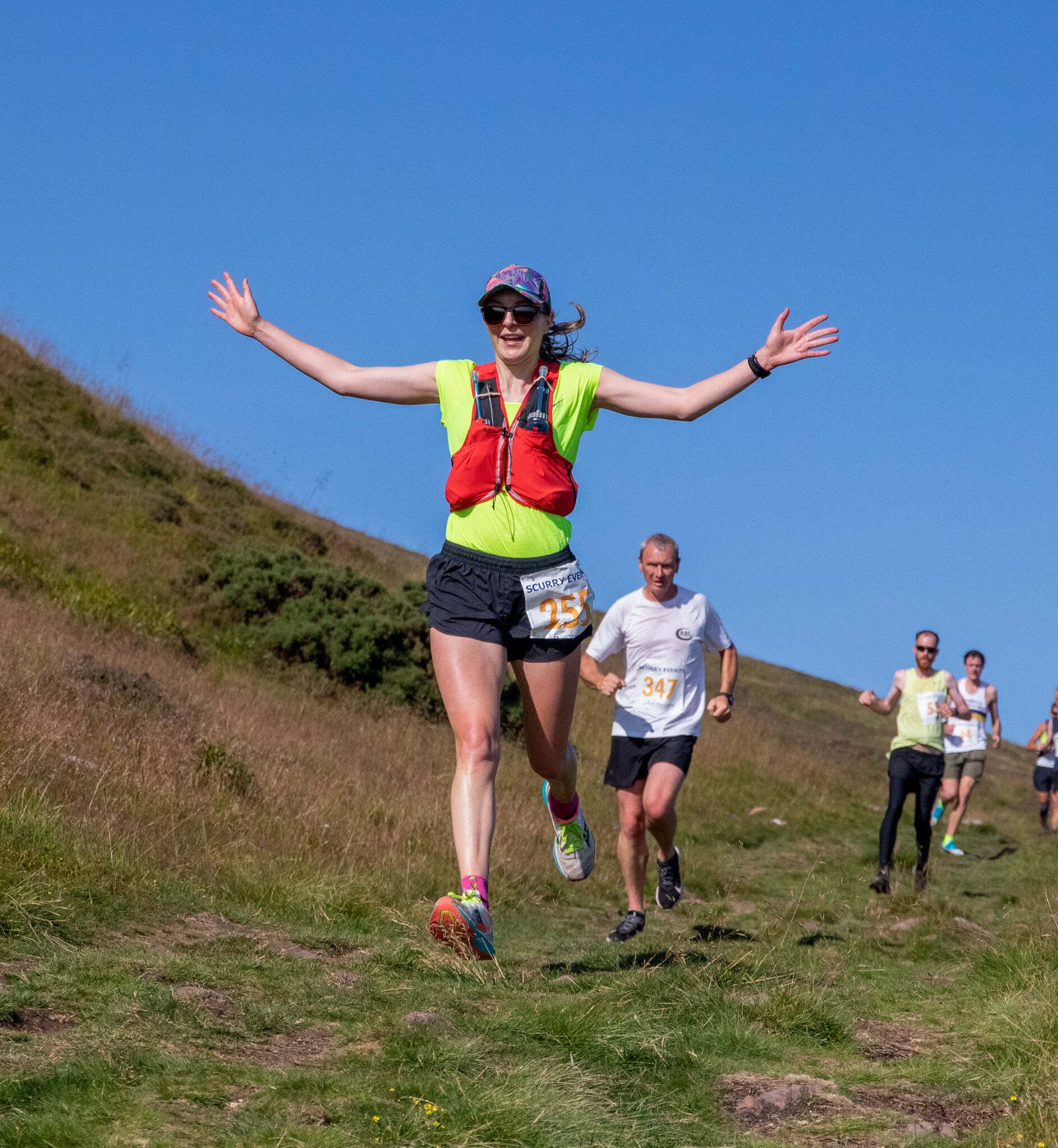 A woman in a neon green shirt and red vest runs joyfully with arms outstretched on a grassy trail during a sunny day. Other runners trail behind her, and the background features a hillside with clear blue skies. She wears sunglasses, a cap, and a race number 273.