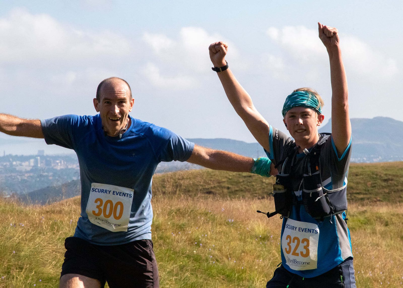 Two runners, both wearing race bibs number 300 and 323, are seen smiling with their arms raised in celebration. They are on a grassy path with hills and a cityscape visible in the background under a partly cloudy sky.