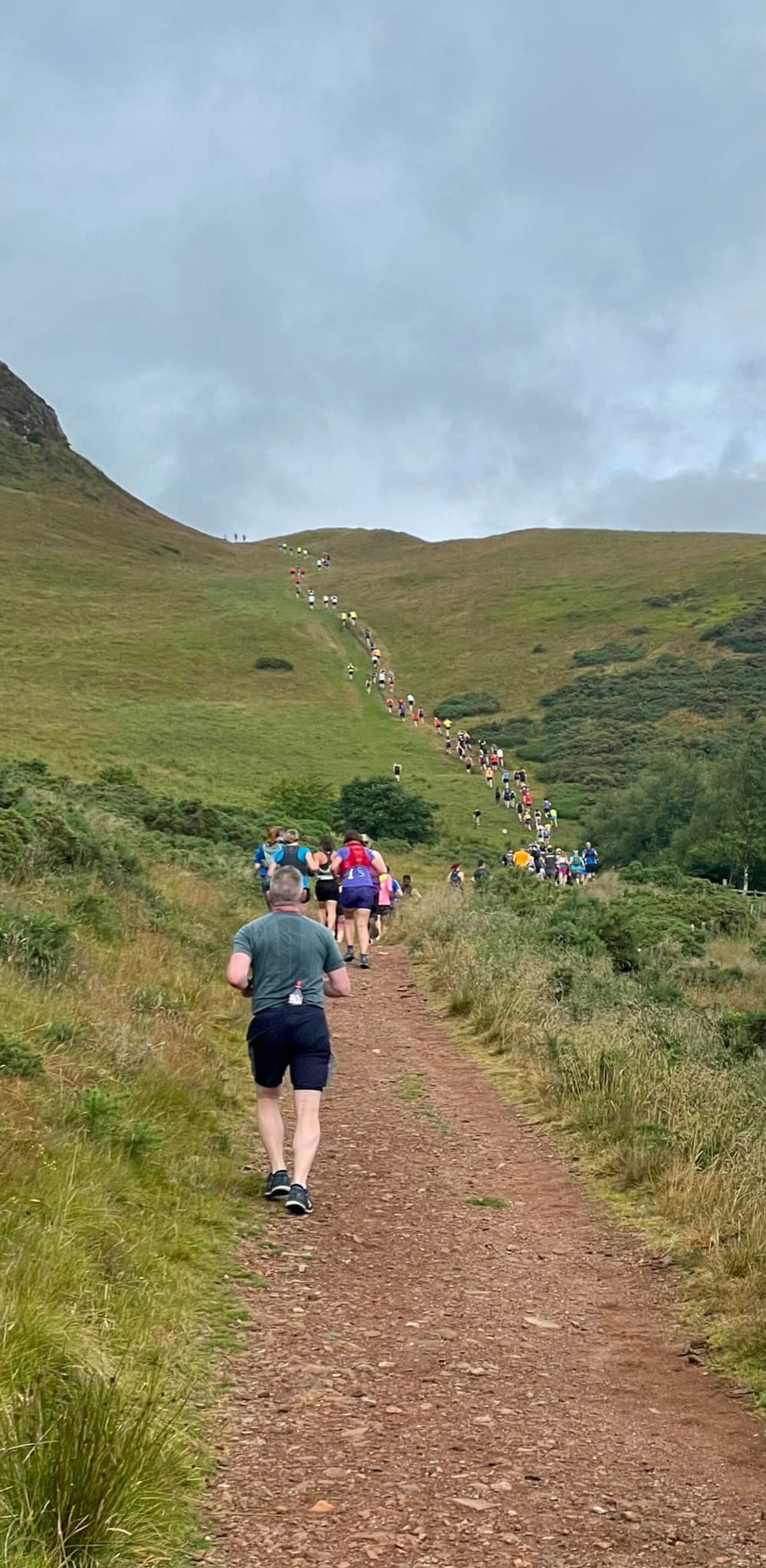 A large group of runners making their way up a hill on a dirt trail surrounded by green grass and shrubs. The sky is cloudy, indicating an overcast day. The participants are wearing various athletic clothing, and the trail winds up towards a steeper incline.