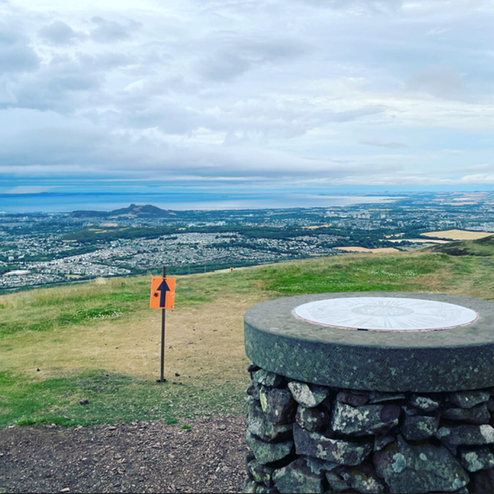 A view from a high vantage point showing a circular stone structure and a direction sign with an arrow. The expansive landscape below includes a town, fields, hills, and a body of water under a cloudy sky.