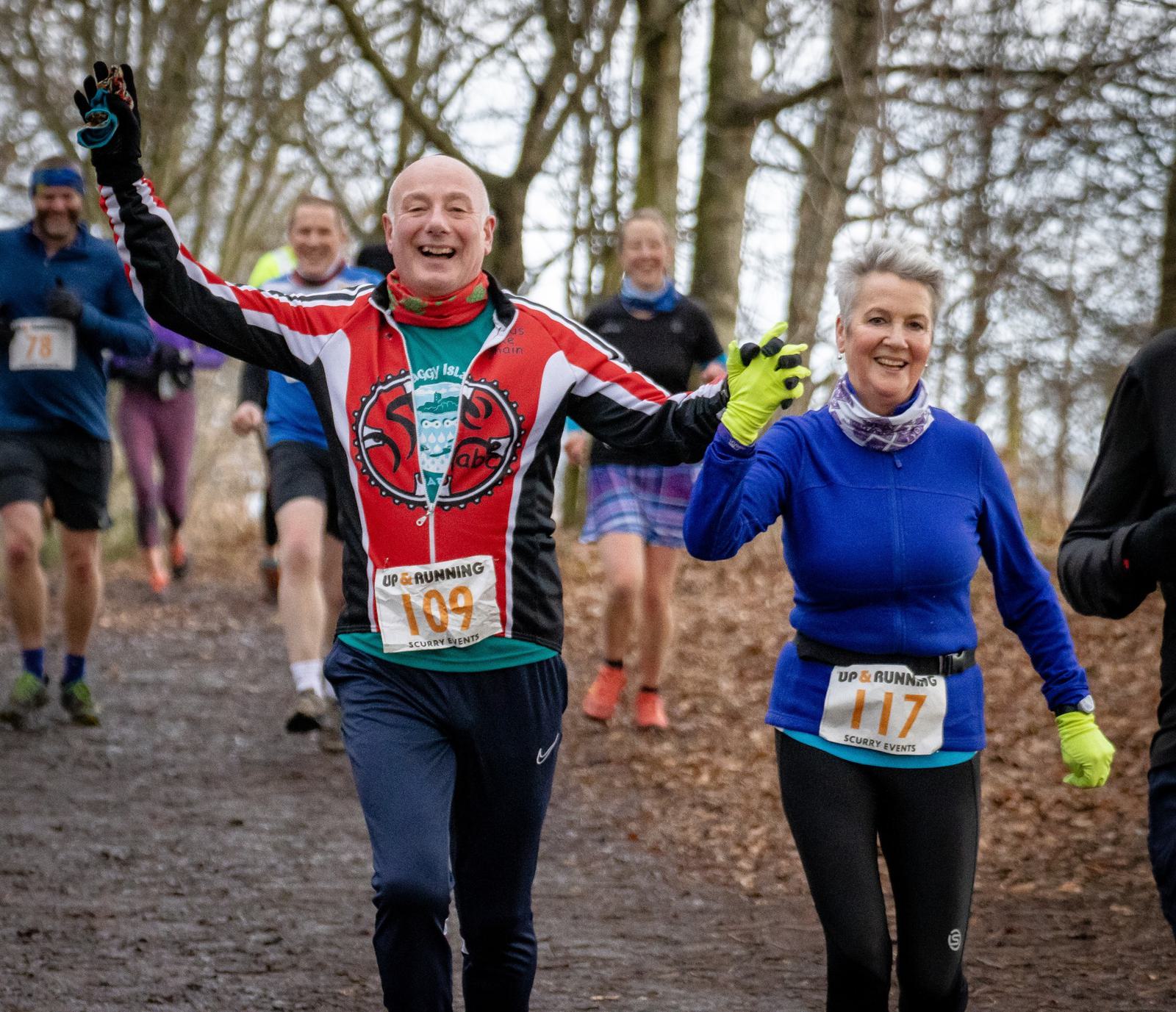 A group of runners participating in a trail race through a forest. Two of them, a man and woman, are in the foreground, smiling broadly and waving at the camera. The man is wearing a red and black jacket with number 109, and the woman is in a blue top with number 117.