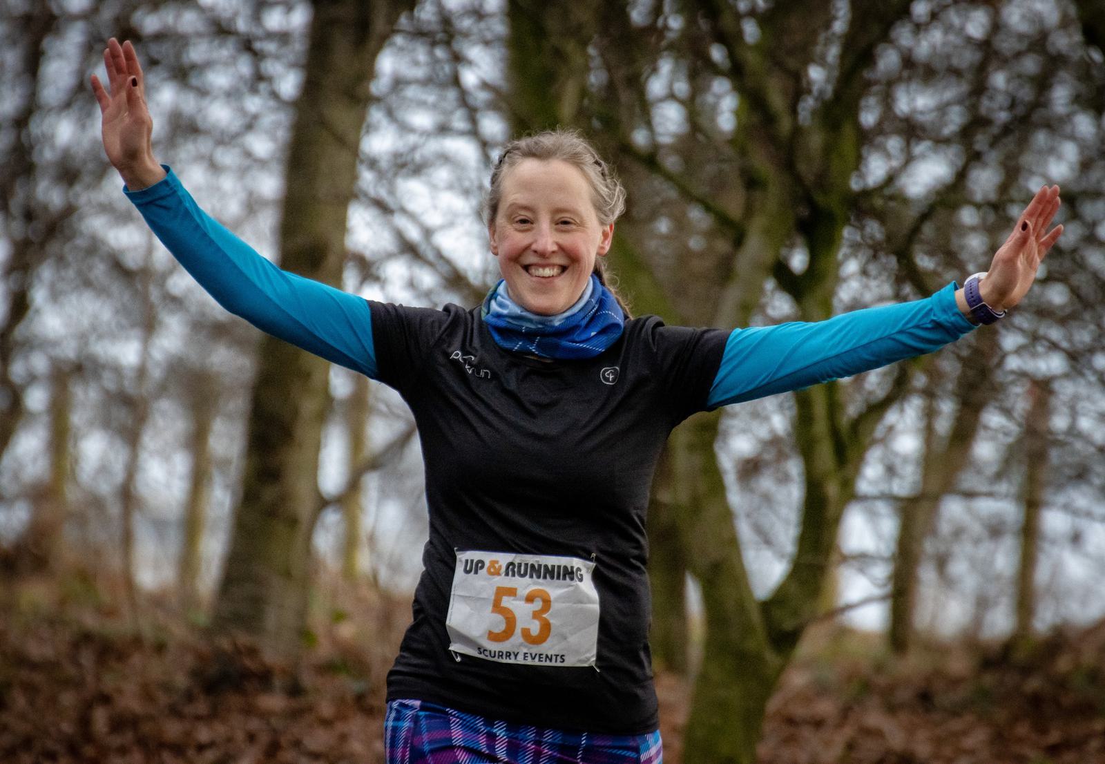 A person in a black shirt and blue sleeves, wearing a race bib numbered 53, smiles and raises their arms in celebration while running in a forested area during a race event. The background features leafless trees.