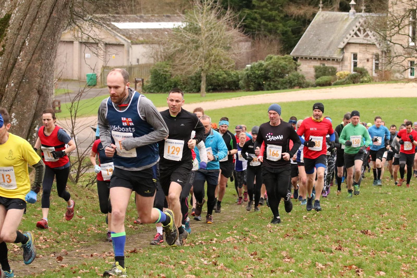 A group of runners, dressed in various athletic gear and numbered bibs, participate in an outdoor race on a grassy path with autumn leaves. Trees and buildings are visible in the background. The weather appears cool, as many runners wear long sleeves and hats.