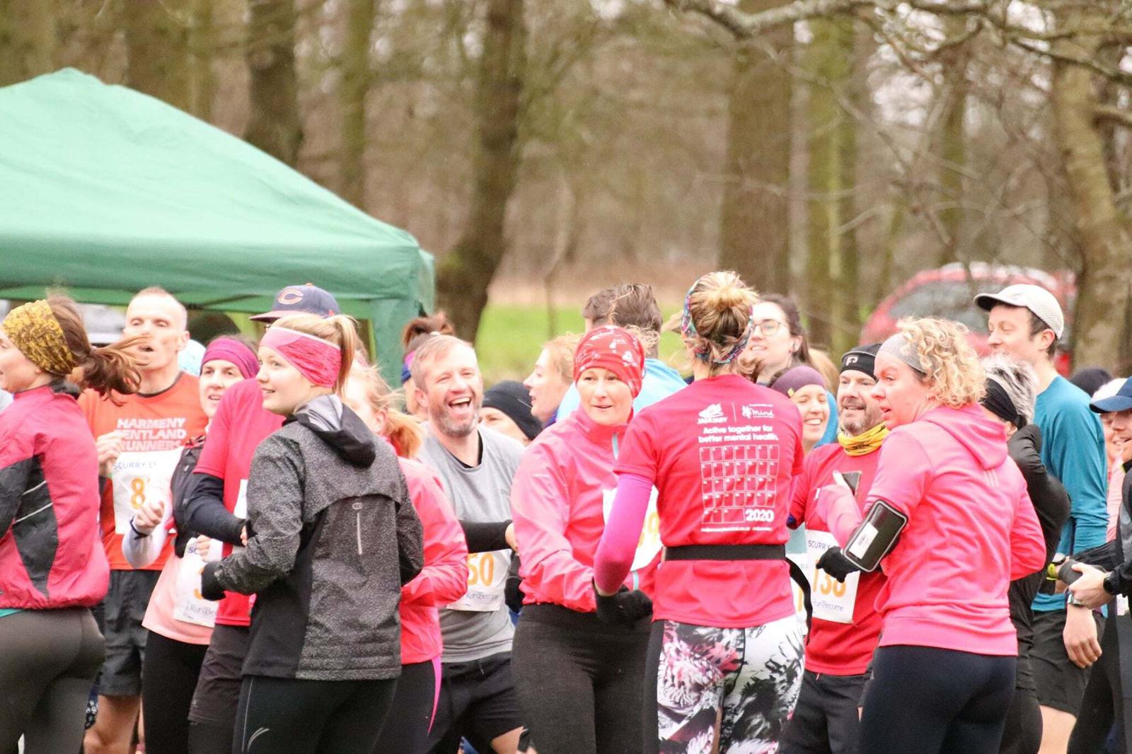 A group of runners stand together in a park area, chatting and laughing before a race. They are dressed in athletic clothing, some wearing red jackets and headbands. A green tent and trees are in the background. The atmosphere appears lively and friendly.