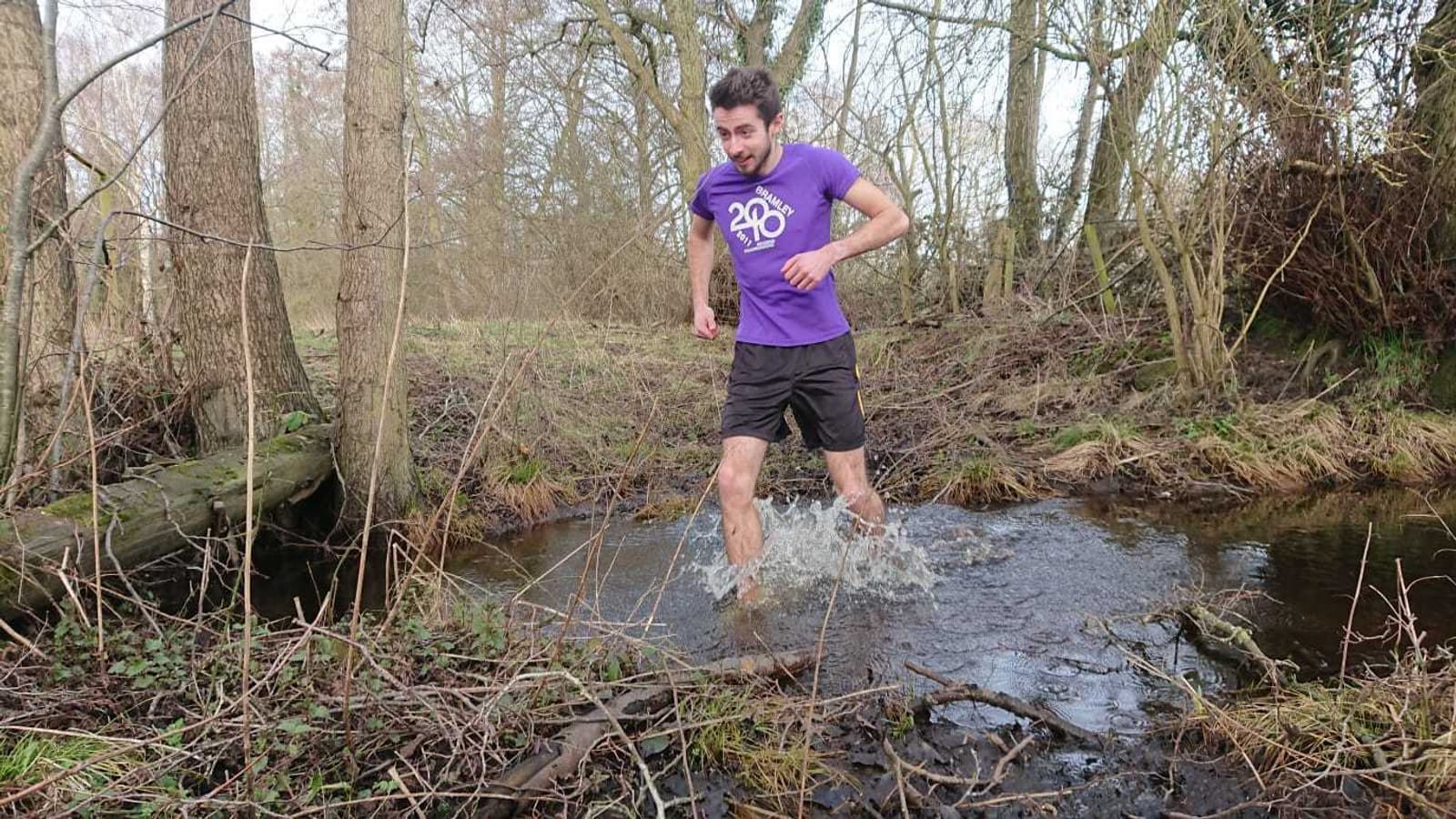 A man in a purple t-shirt and black shorts is running through a shallow stream in a forest. He is making a splash with his feet as he crosses the water. The area around him is filled with leafless trees and brown, grassy terrain.