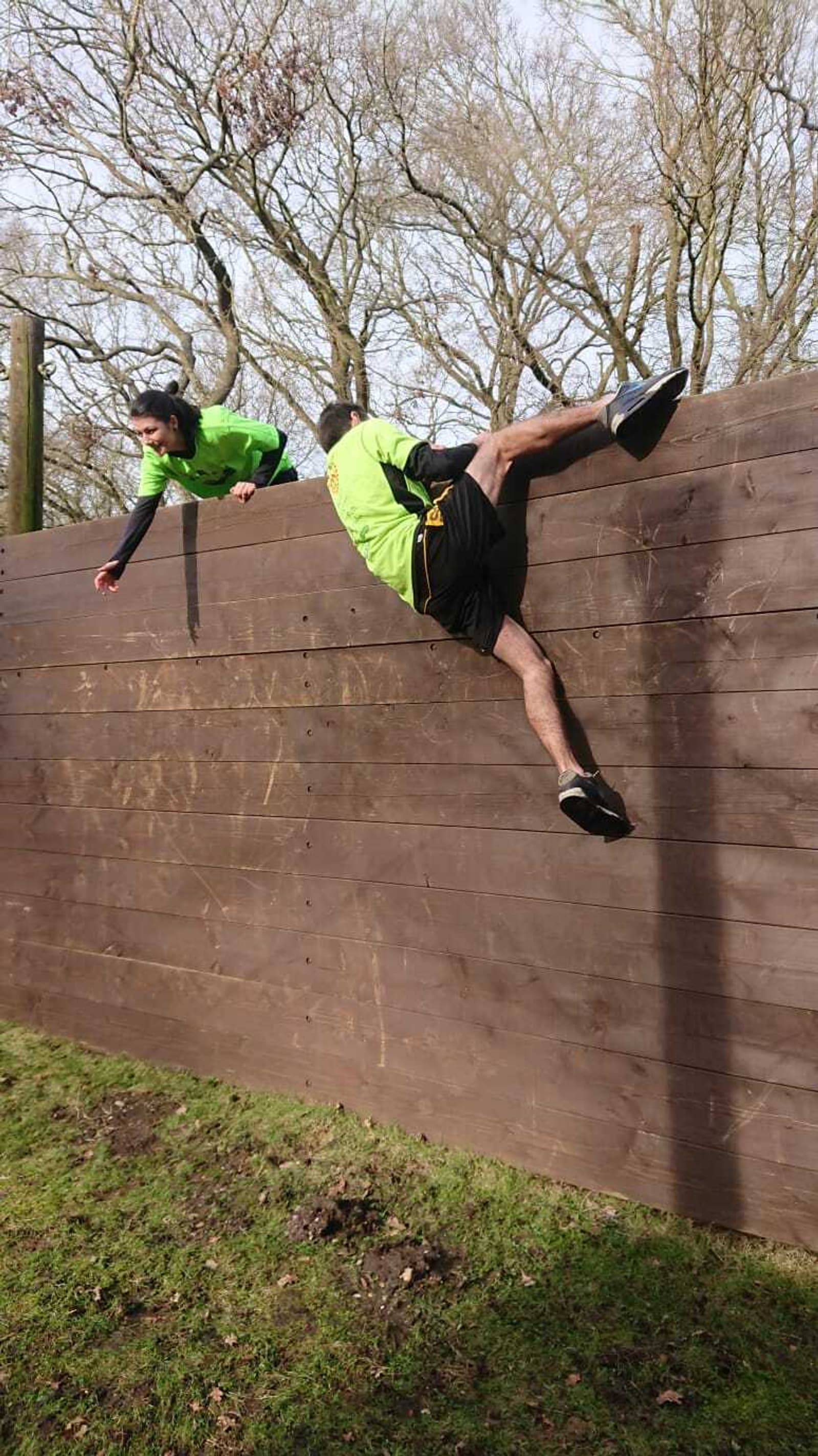 Two people in green shirts are participating in an outdoor obstacle course. One person is helping the other over a tall wooden wall. The second person has one leg over the wall, and their hands are gripping the top as they pull themselves up. Trees are visible in the background.