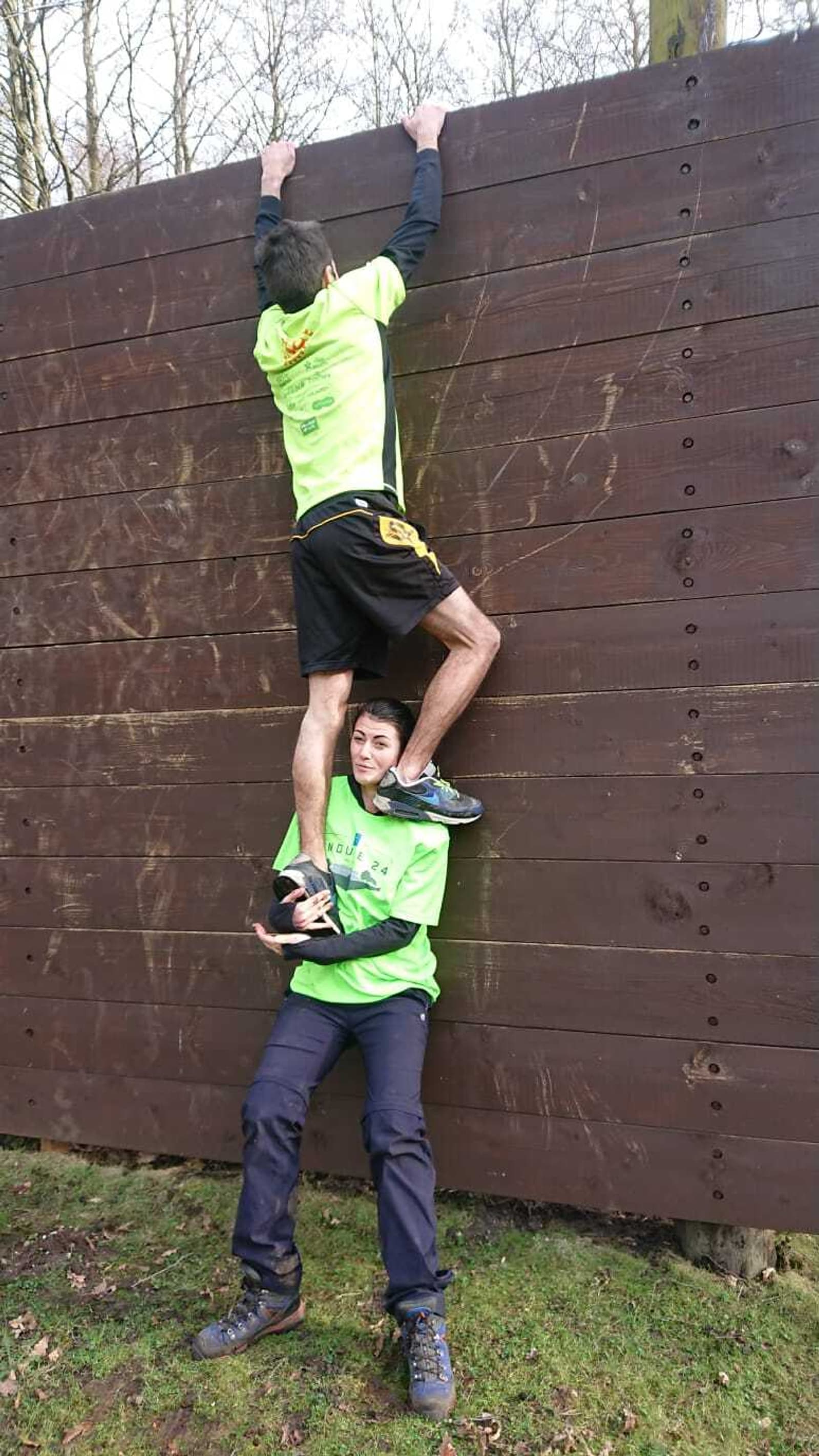 Two individuals wearing neon green shirts are attempting to scale a tall wooden wall outdoors. One person, supported by the other, has their hands gripping the top of the wall, while the other person is holding them up from below, standing on grass.