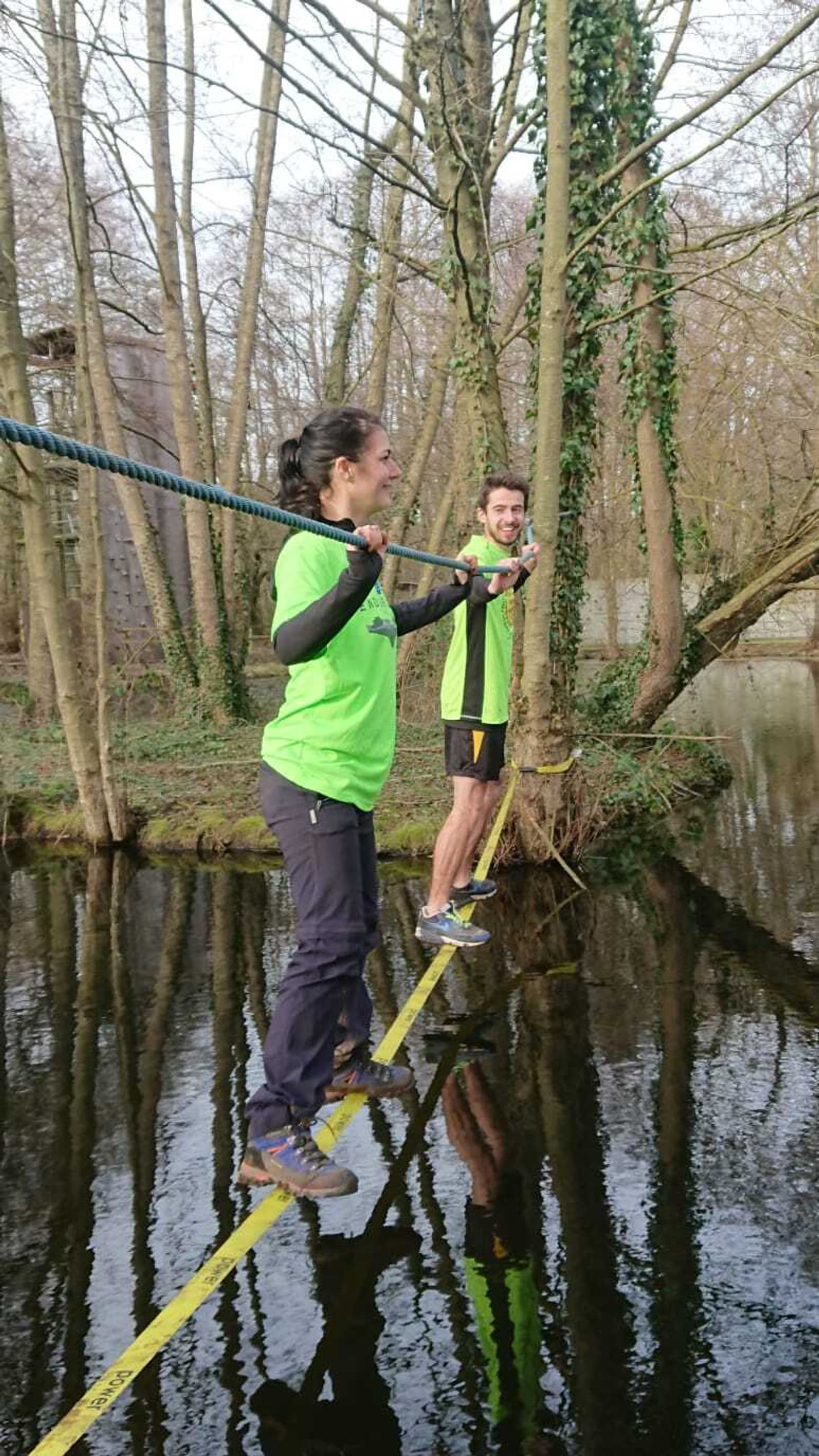 Two people balancing on a slackline above water in a wooded area. They are wearing bright green shirts and holding a rope for support as they traverse the line. The trees around are tall and leafless, suggesting a leafless season. Both individuals look focused.