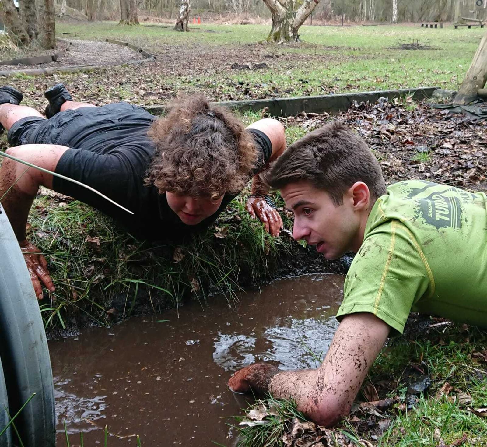 Two people are crawling in a muddy obstacle course with half their bodies submerged in a trench filled with muddy water. They are both covered in mud and appear focused on the task. The surrounding area is grassy with some trees in the background.