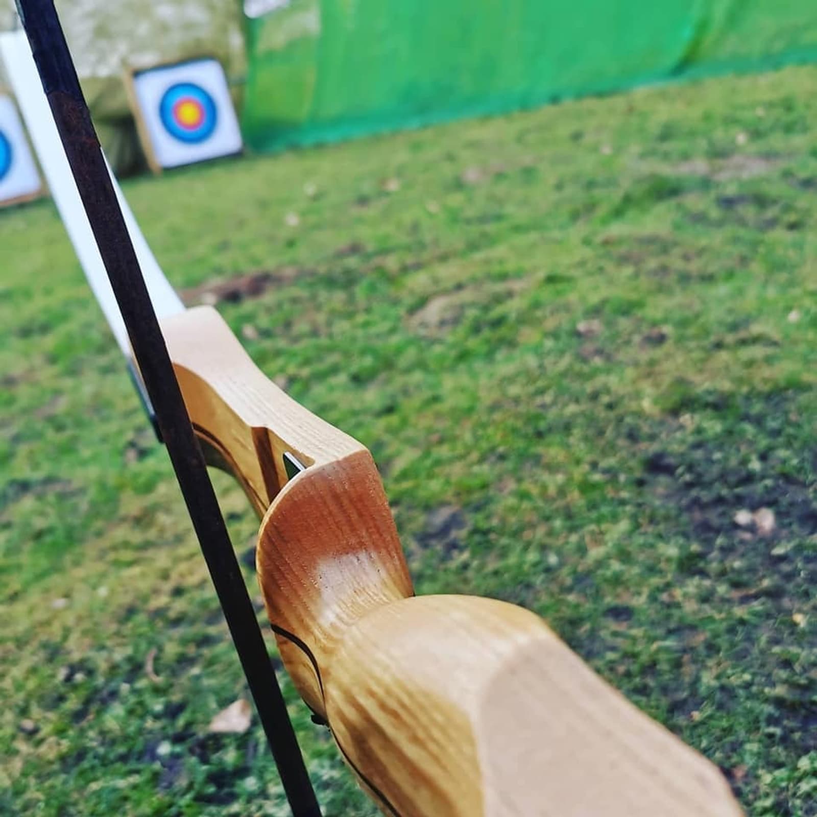 Close-up shot of a wooden archery bow with a string in focus. Two target boards with multicolored bullseyes are in the blurred background, positioned on a grassy field. The scene appears to be an outdoor archery range.