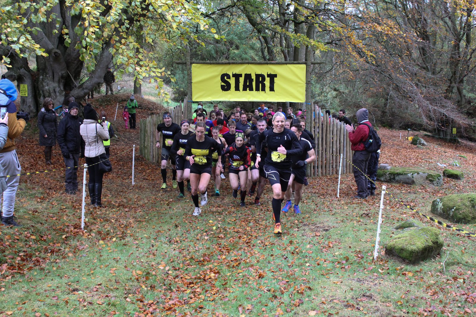Participants in sports attire are running through a "START" arch at the beginning of a trail race in an autumnal forest setting. Spectators and trees with yellow leaves are visible in the background. The ground is covered with fallen leaves and patches of green grass.